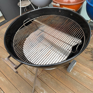 A black round charcoal grill with a stainless steel grate and a round metal tray, shown on a wooden deck with other grills in the background.