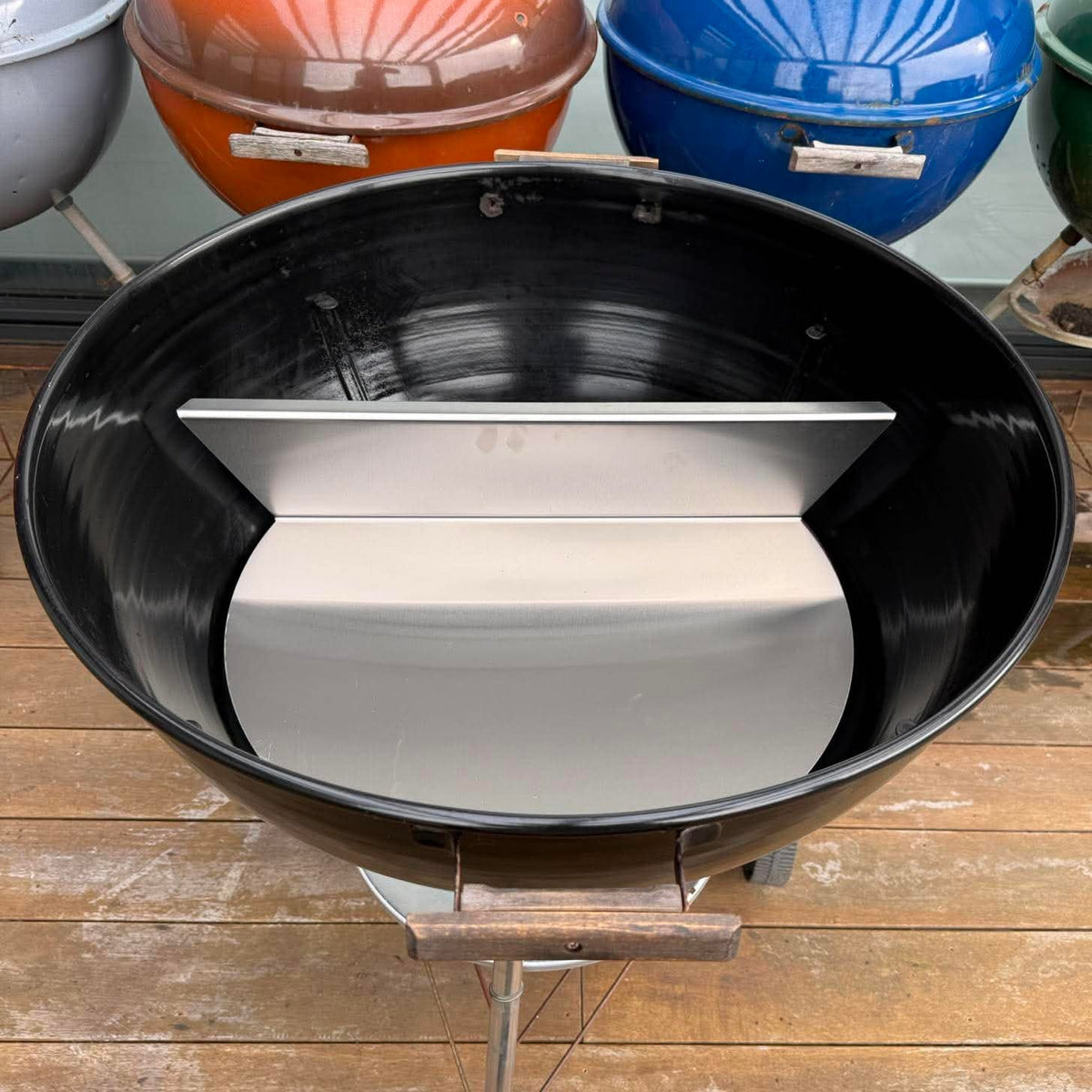 A black round charcoal grill with a silver metal divider tray inside, set on a wooden deck. Other colorful grill domes are visible in the background.