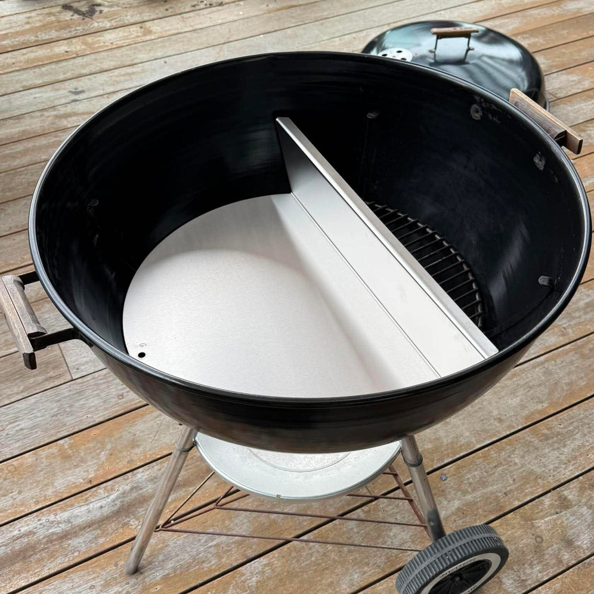 A black round charcoal grill with its lid off, sitting on a weathered wooden deck. A white circular insert or liner is visible inside the grill, along with a metal divider plate and the silver metal legs and wheel.