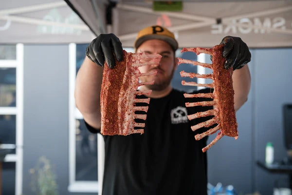 Pitmaster holding seasoned racks of ribs ready for the smoker at Smoke School BBQ class